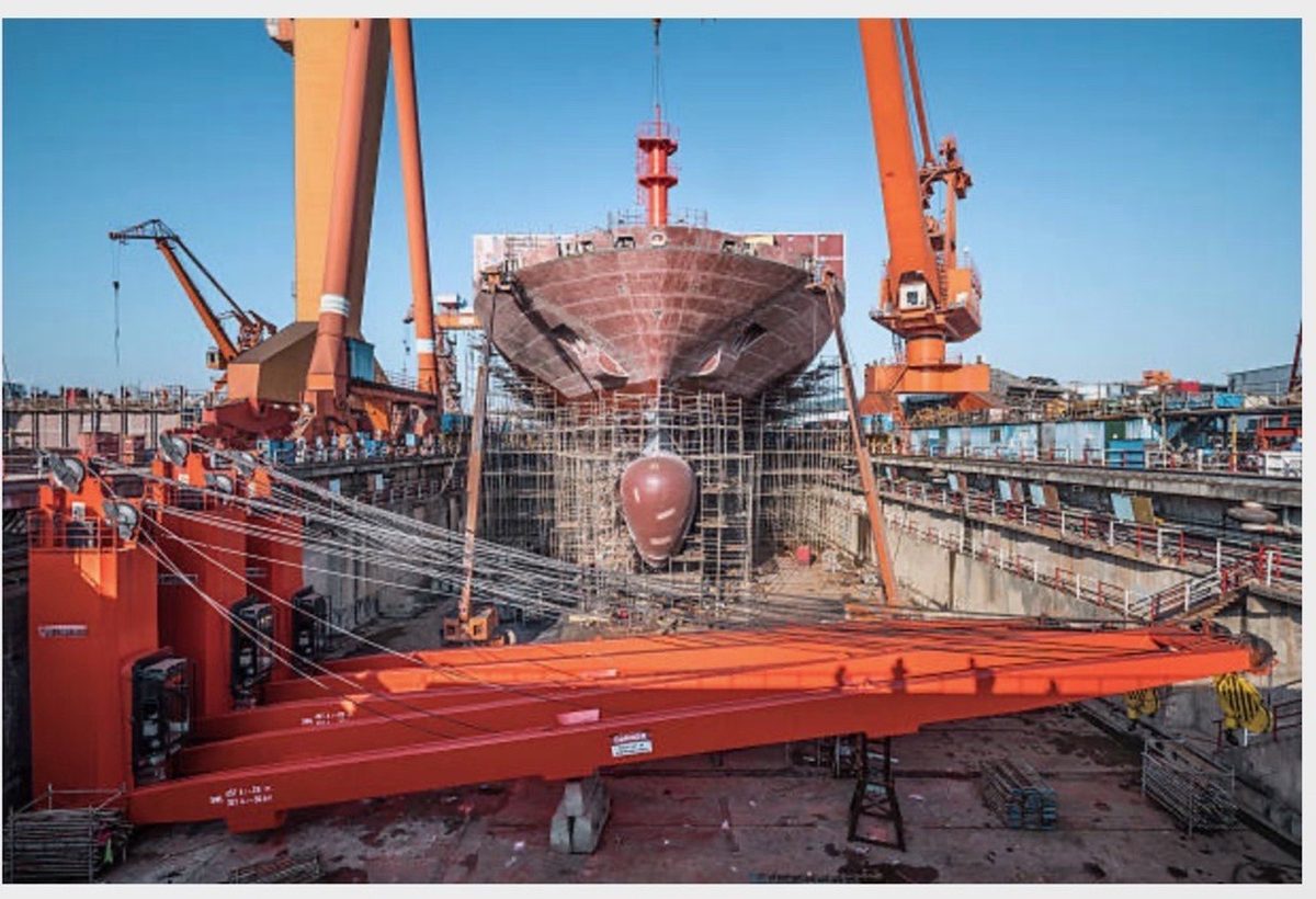 Ship's hull under construction in a shipyard dry dock with gantry cranes overhead